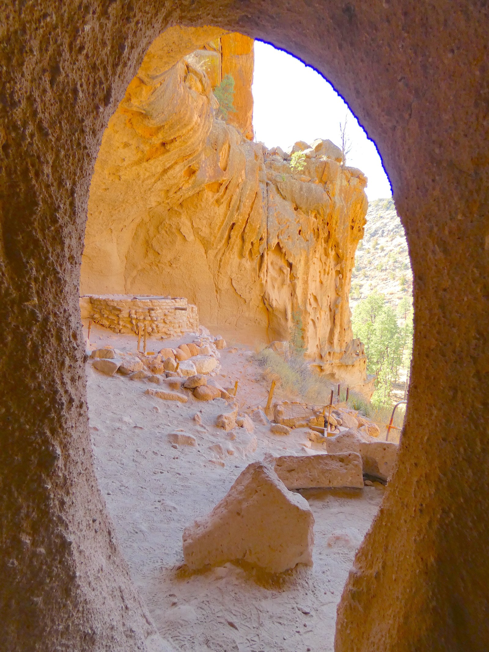 Looking out from the shadowy interior of a large cavate (Alcove House) at Bandelier National Monument, framing a view of a reconstructed kiva, stone ruins, and the sunny cliff face outside