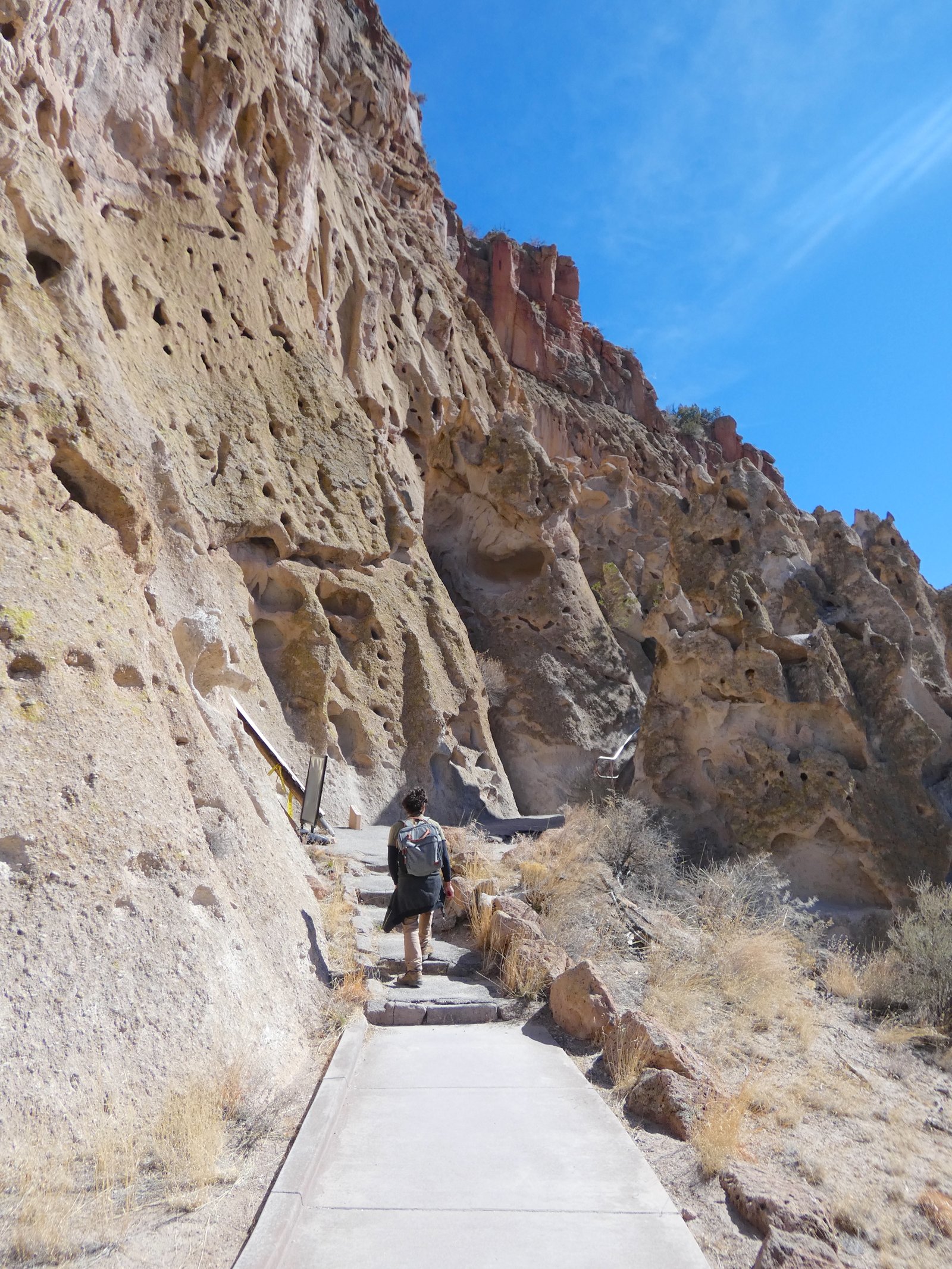 A hiker wearing a backpack and hiking boots on the sunny Main Trail right next to the towering volcanic cliff at Bandelier National Monument.