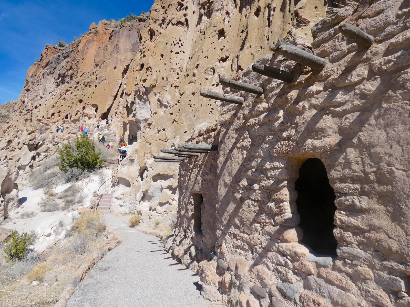 Hikers walking along the paved Main Loop Trail passing the historic stone ruins of Long House at Bandelier National Monument.