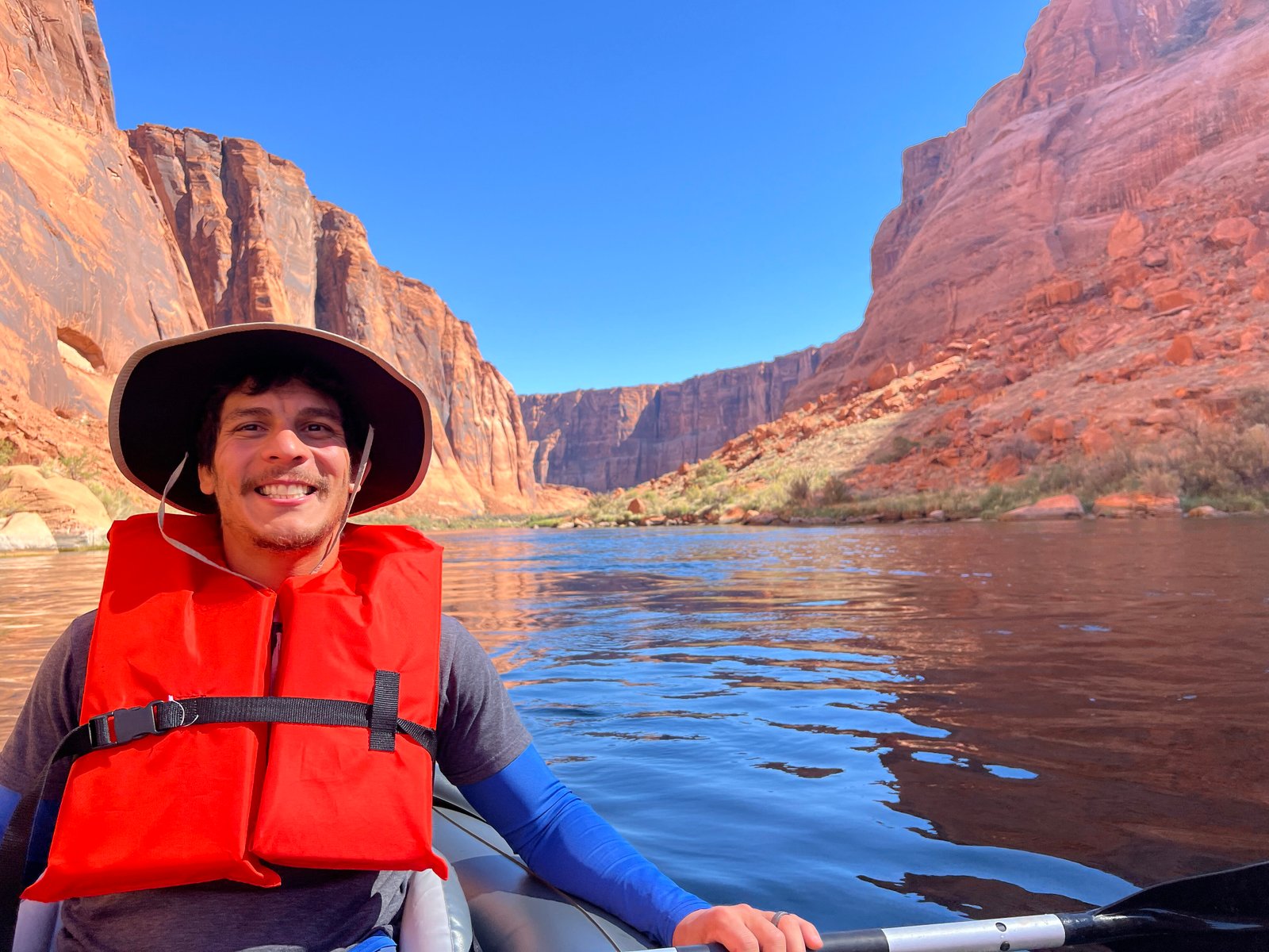 person wearing hat and life jacket sitting in kayak in the river with red canyon walls behind