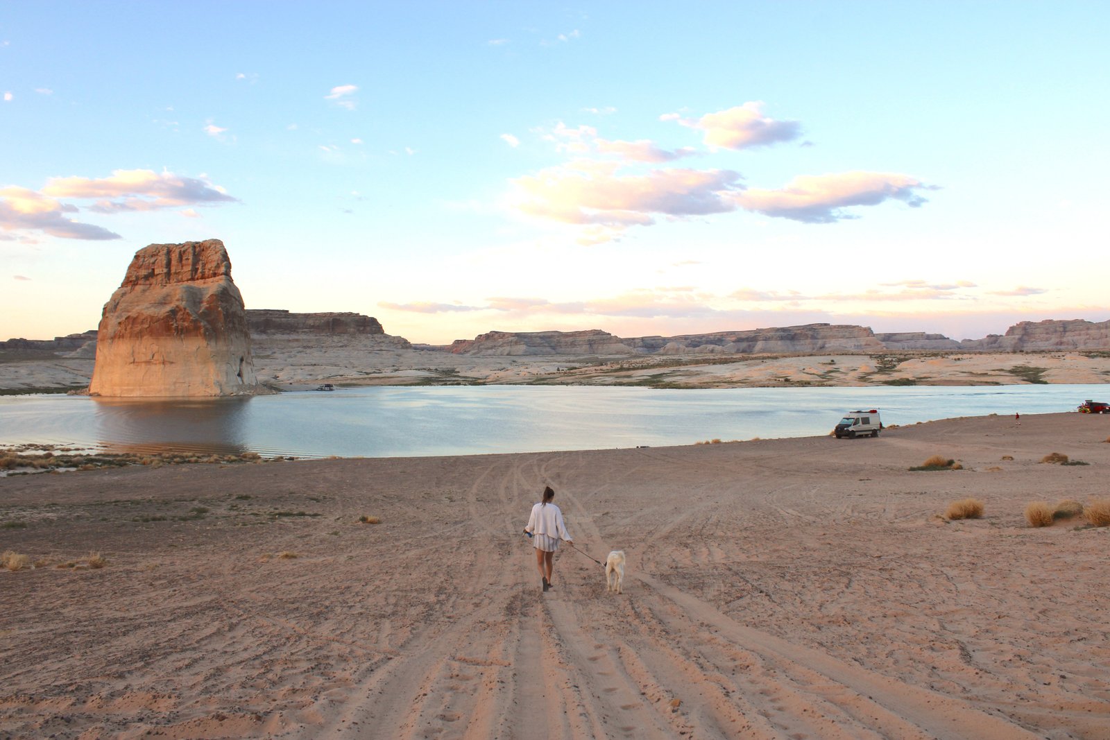 person and dog walking on sand with lake and large rock in the distance