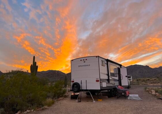 Travel trailer parked in an Arizona desert campground at sunset with saguaros and mountains.