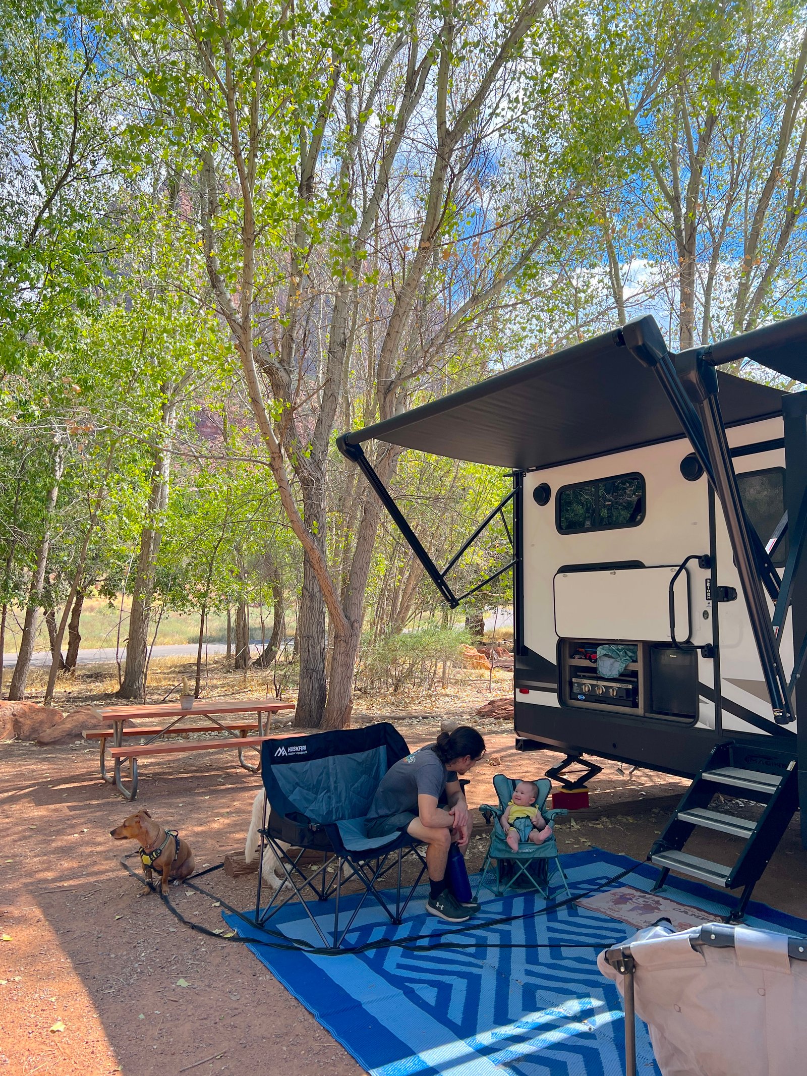 Father and toddler daughter relaxing in camping chairs at their campsite in Watchman Campground with Zion landscape in the background.