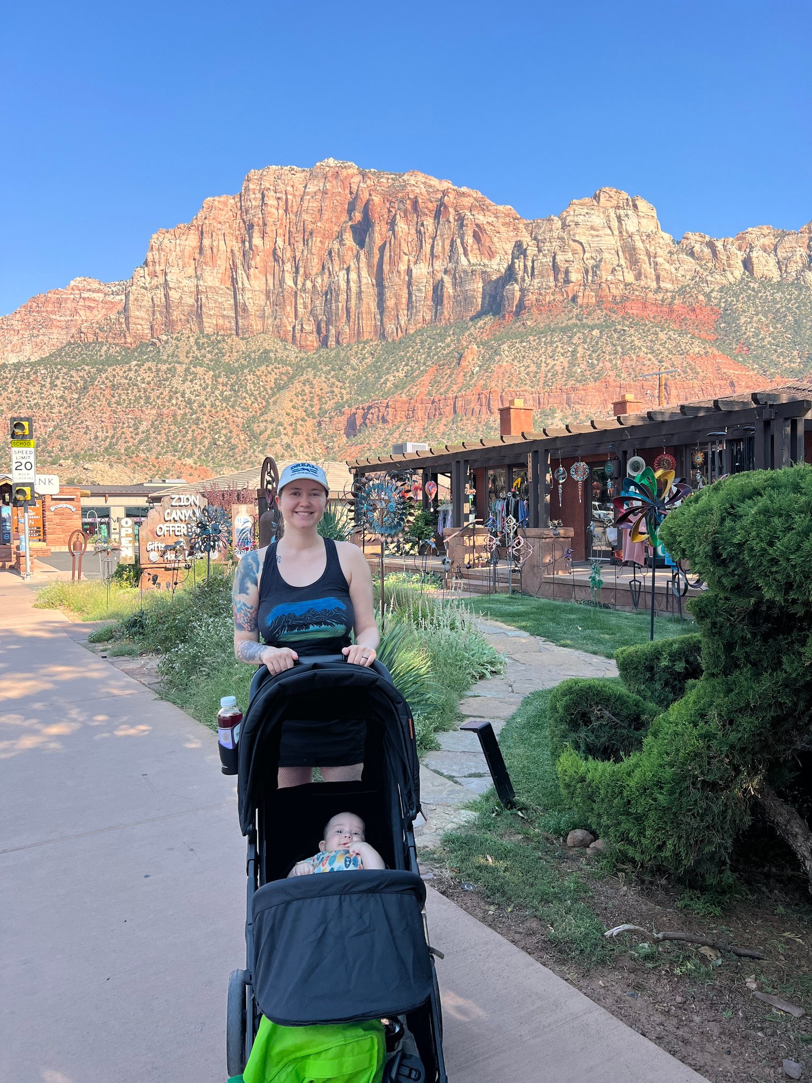 Mother pushing a toddler daughter in a stroller down the sidewalk in the town of Springdale, Utah, with Zion Canyon visible in the distance.