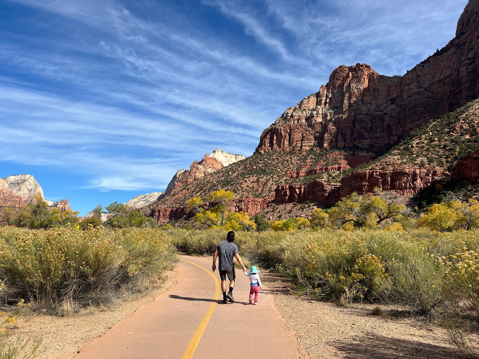 Father and toddler daughter walking together on the paved Pa'rus Trail with towering Zion Canyon walls in the background.
