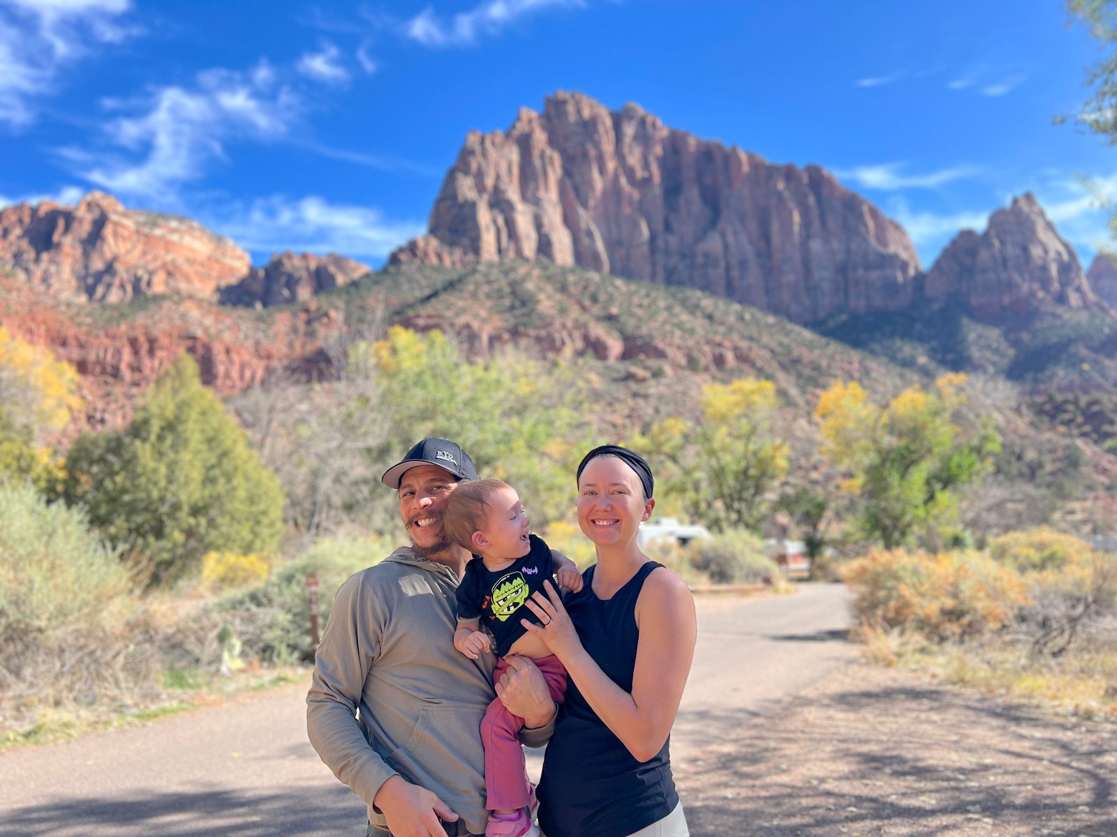 Happy family of three—mother, father, and toddler daughter—posing together on a trail with sweeping views of Zion Canyon and autumn foliage in the background.