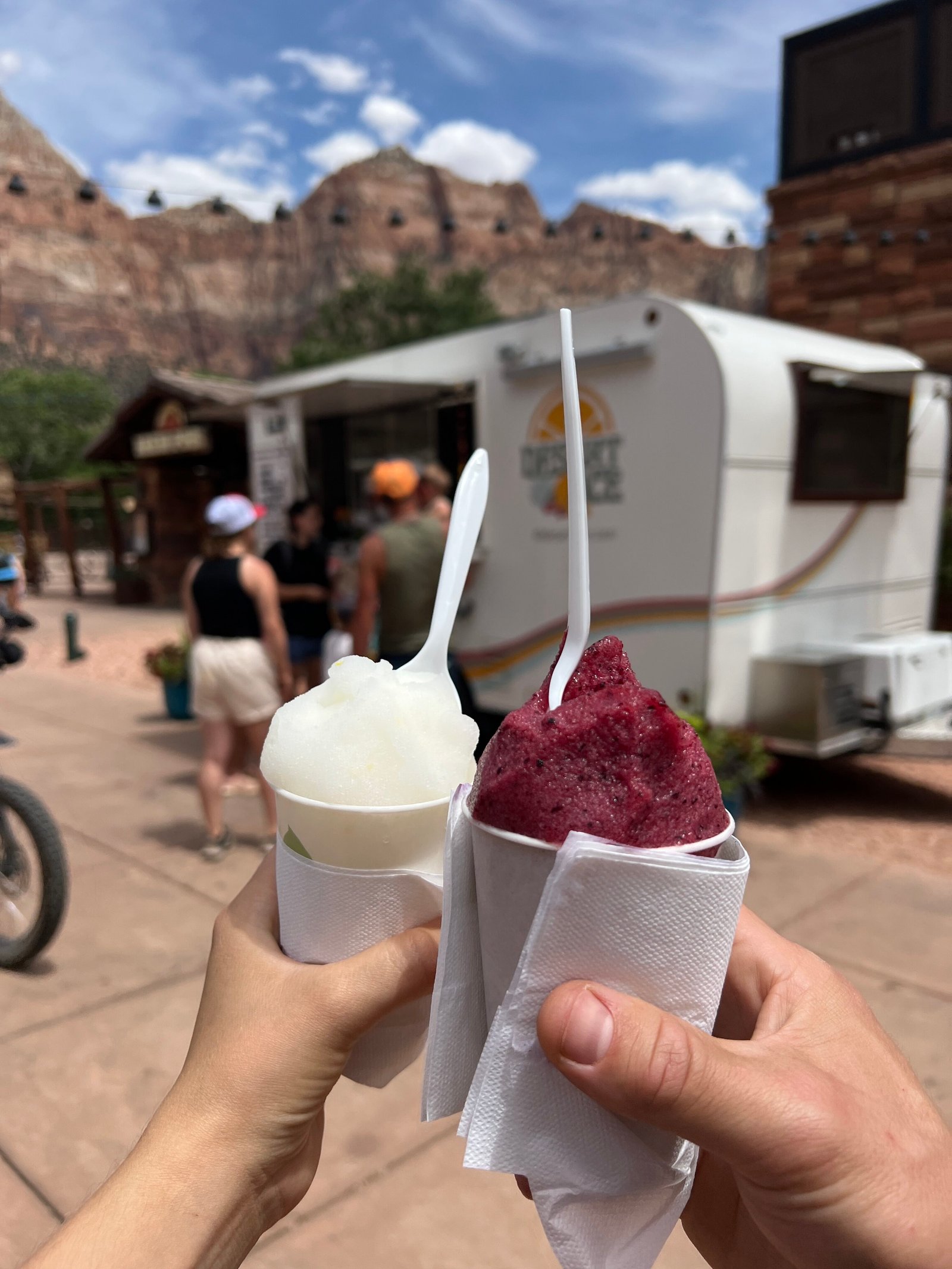 Close-up of two hands holding colorful ice treats (iceys) with the buildings of Springdale and distant Zion rock formations in the background.