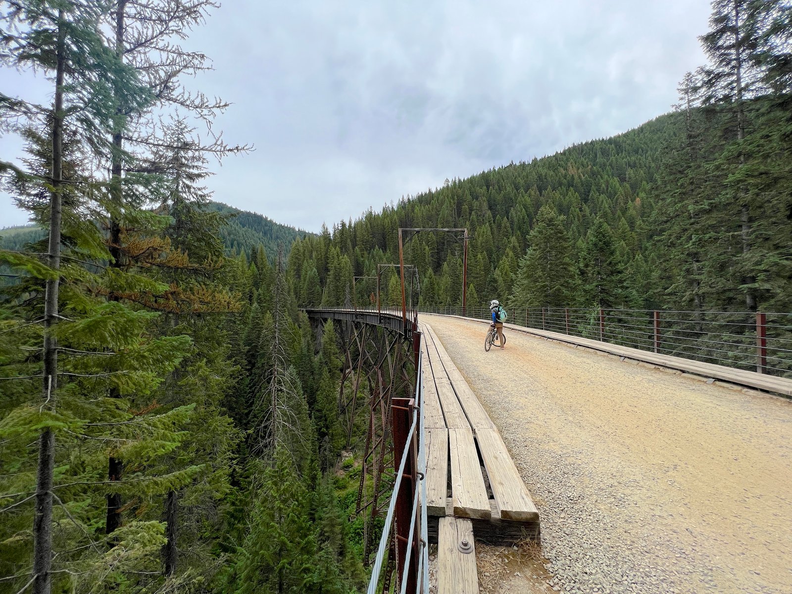 Wide-angle view of a cyclist on a bicycle crossing a tall wooden trestle bridge surrounded by evergreen mountains on the Route of the Hiawatha trail in Idaho.