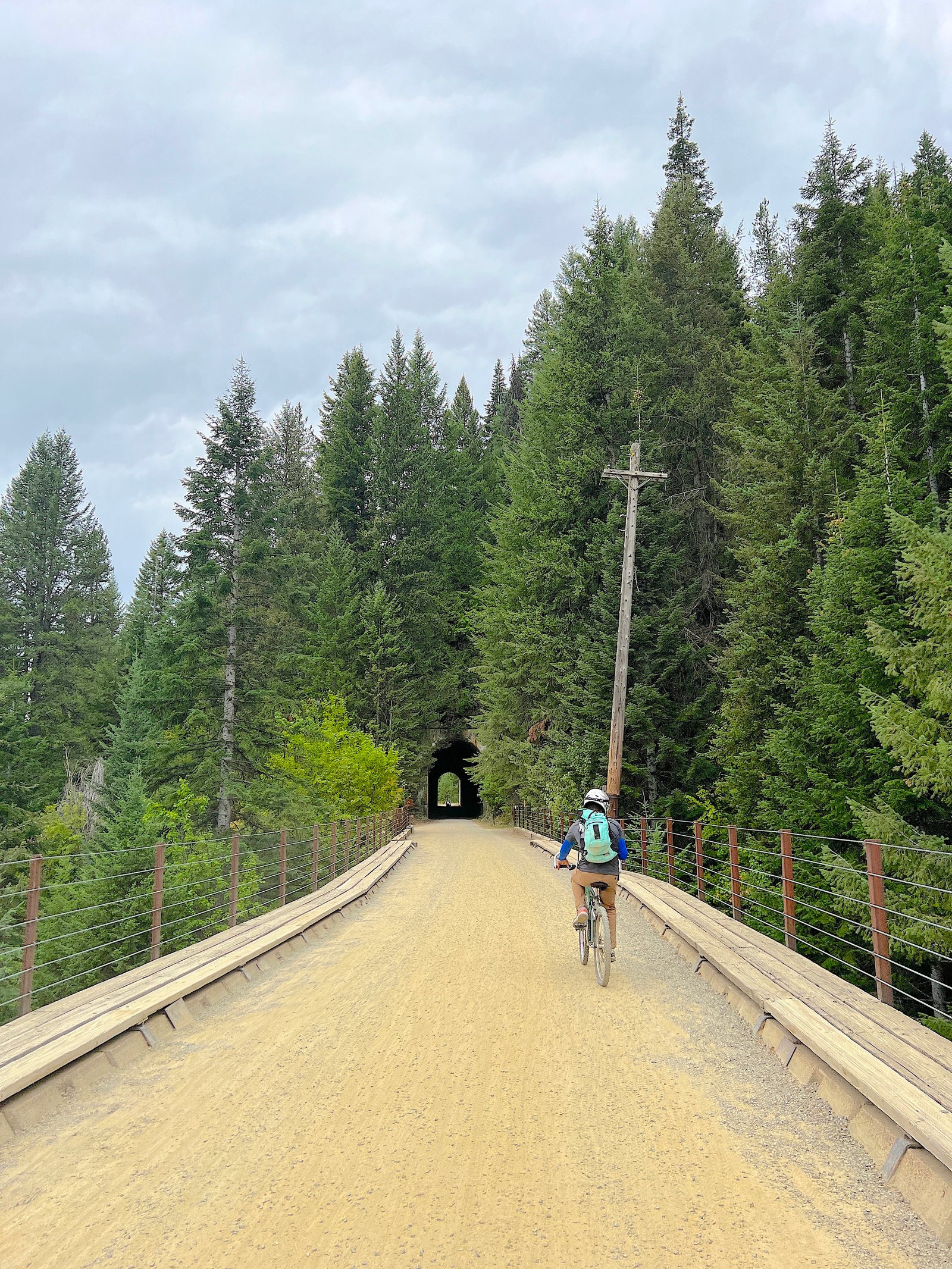 Cyclist riding a bicycle along a dirt rail-trail toward the entrance of a dark mountain tunnel on the Route of the Hiawatha.