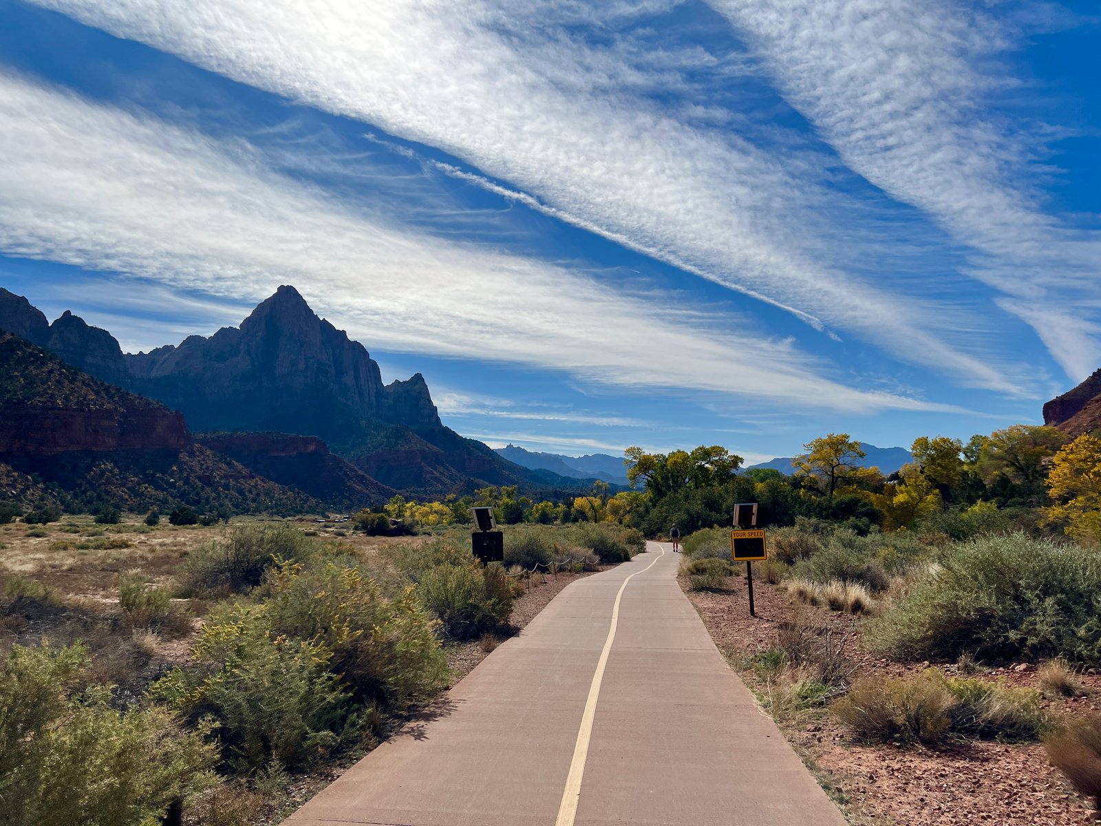 A paved walking and biking path winds through a valley with low shrubs, yellow autumn trees, and a towering mountain peak under a blue sky streaked with long clouds.
