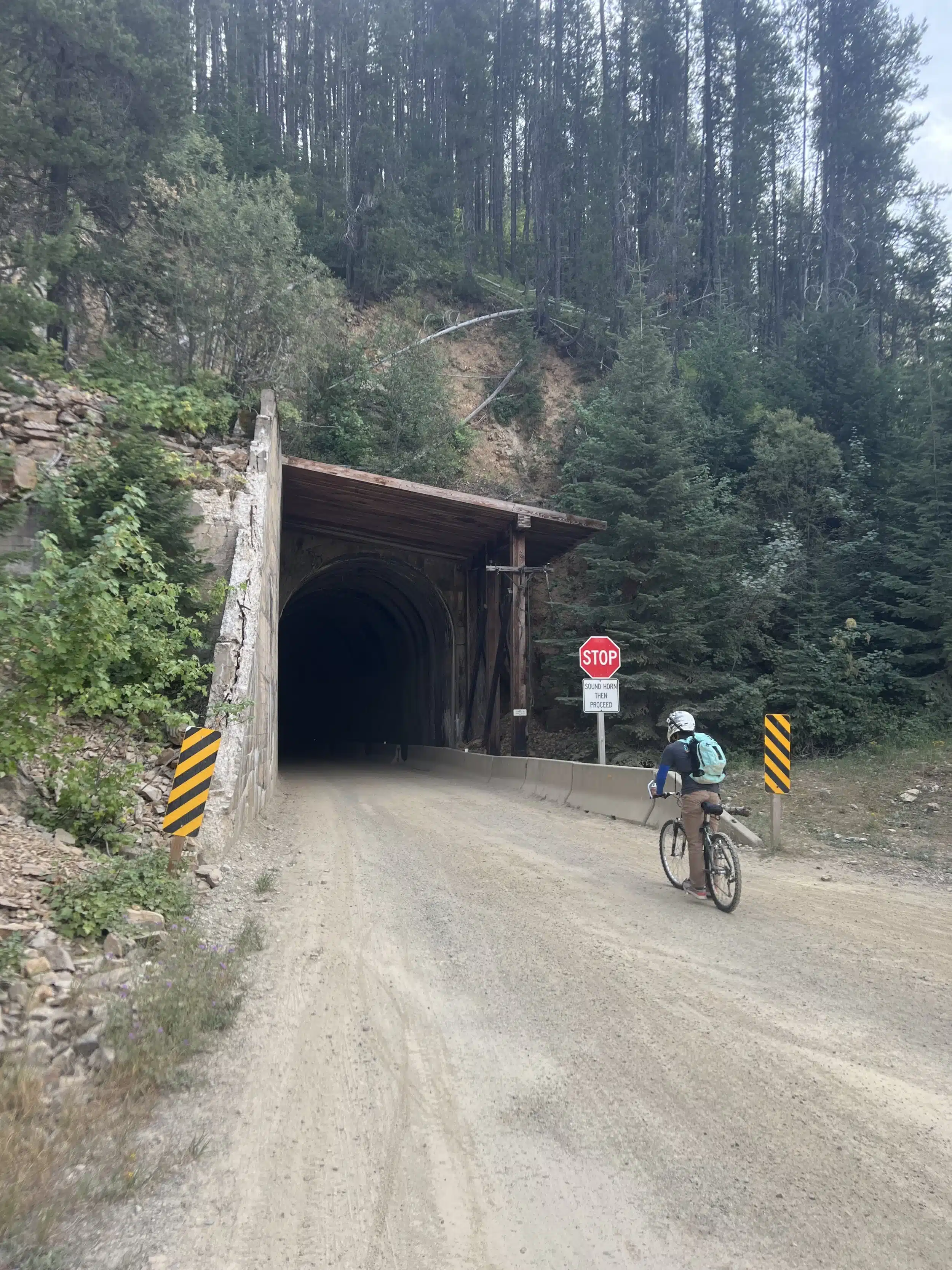 A man on a mountain bike riding along a dirt path toward the dark, shadowed entrance of a tunnel on the Route of the Hiawatha.