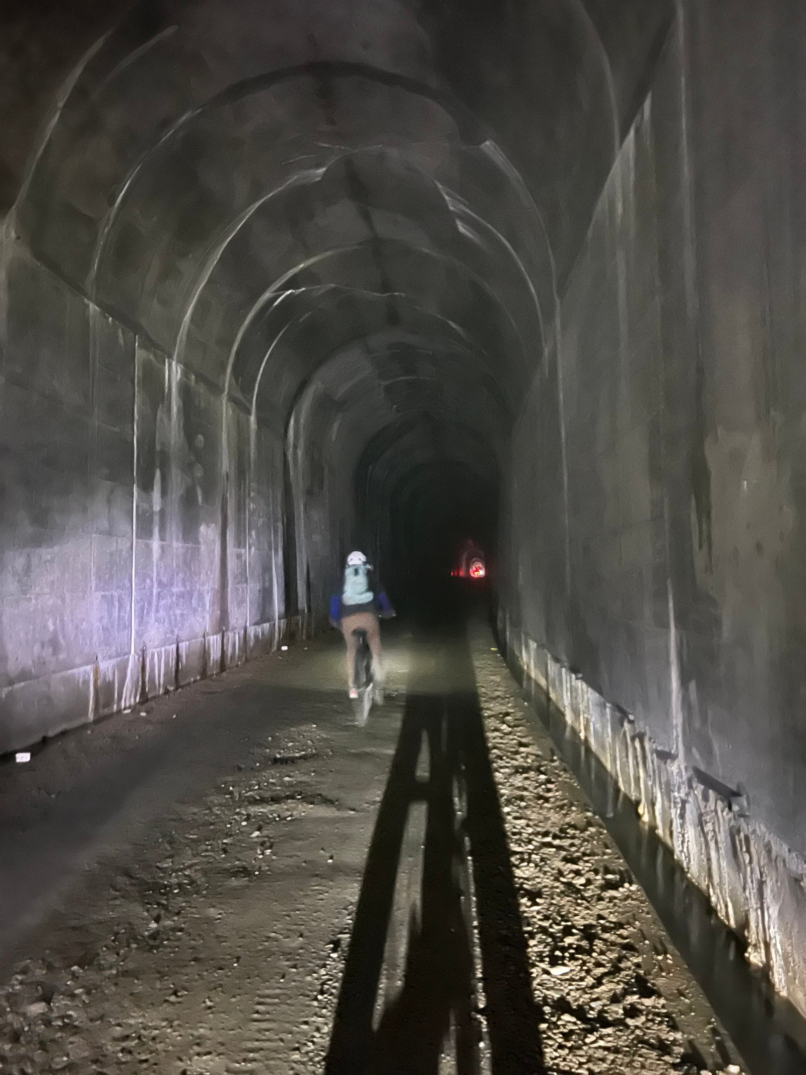 Bicyclist riding through the pitch-black, dark interior of the Taft Tunnel with a bike light illuminating the gravel path on the Route of the Hiawatha.
