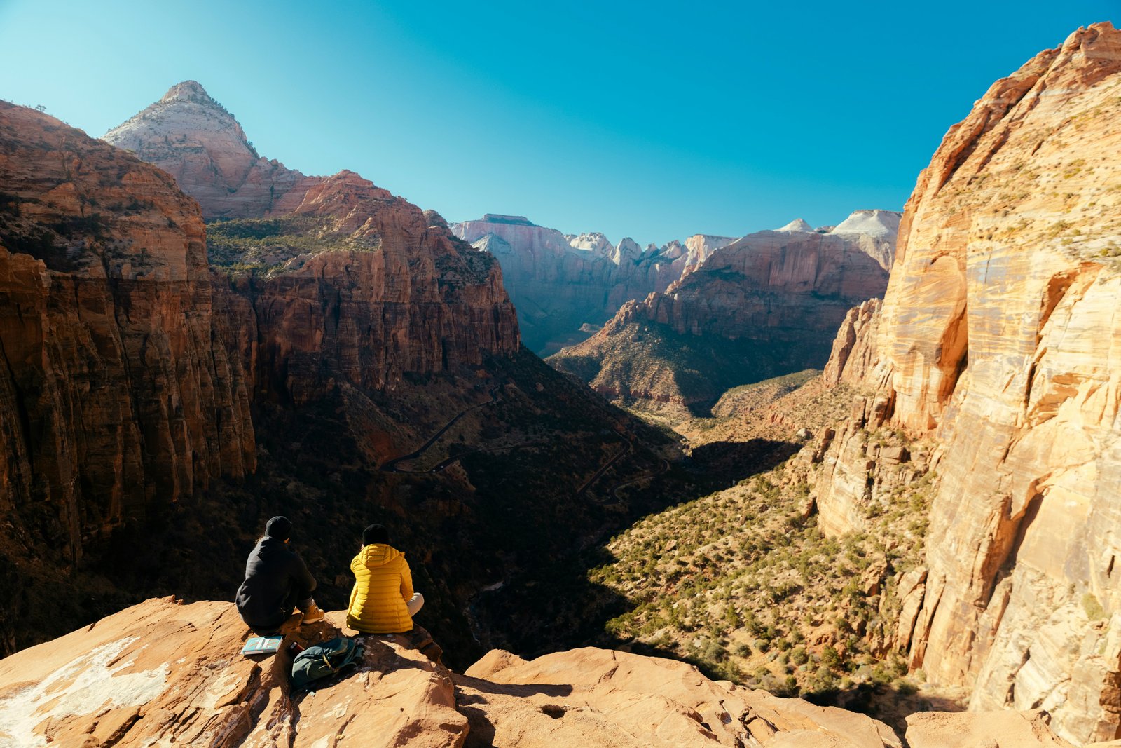 Two hikers sitting on a rocky overlook, facing away from the camera and taking in a vast, panoramic view of a deep desert canyon and mountain peaks under a clear blue sky.