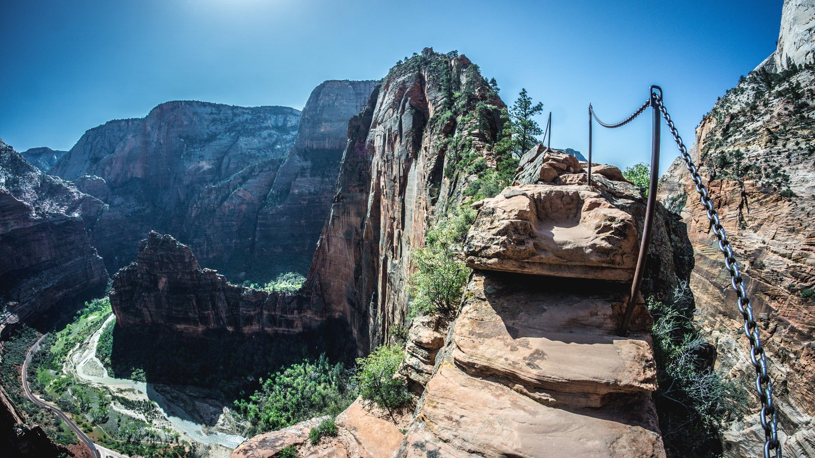 A narrow, rocky trail along the ridge of Angels Landing in Zion National Park, featuring safety chains on the side and a steep drop-off overlooking the canyon and river below