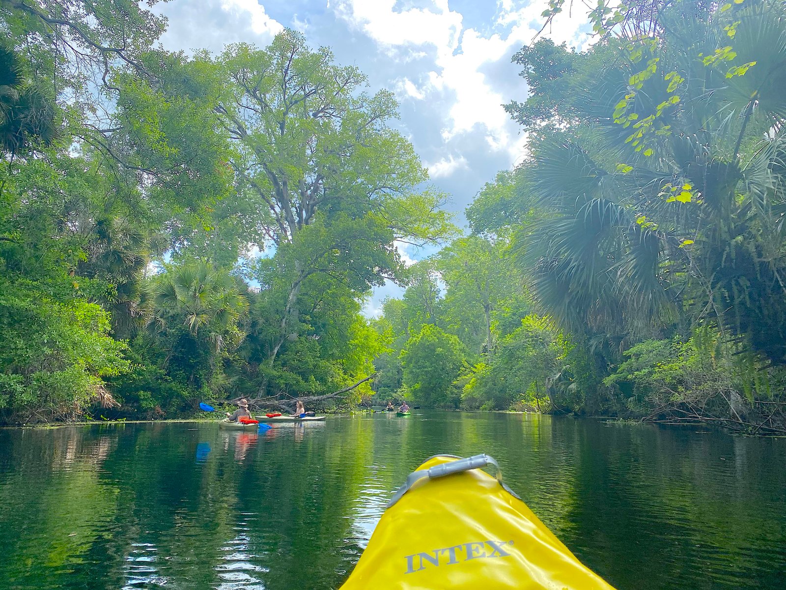 First-person view from the yellow bow of an inflatable kayak paddling down a calm, sunlit river surrounded by lush green trees and palms at Wekiwa Springs State Park.